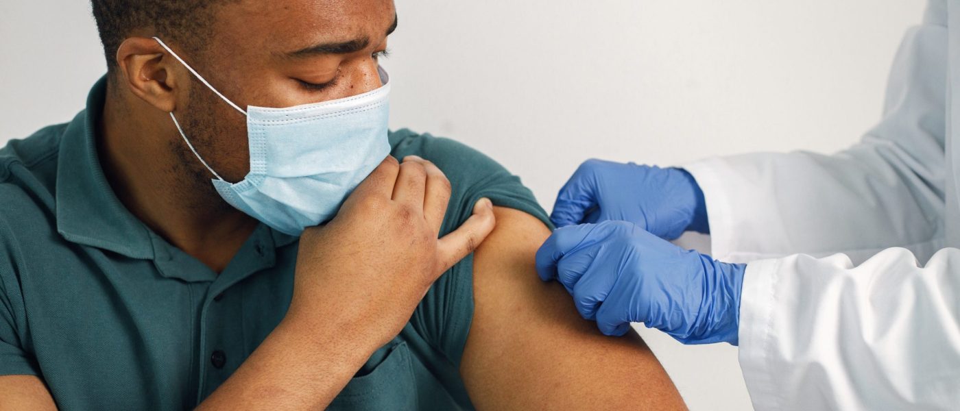 Black man sitting isolated on a white background. Doctor in medical gown stick a band-aid. Man wearing blue shirt and medical face mask.